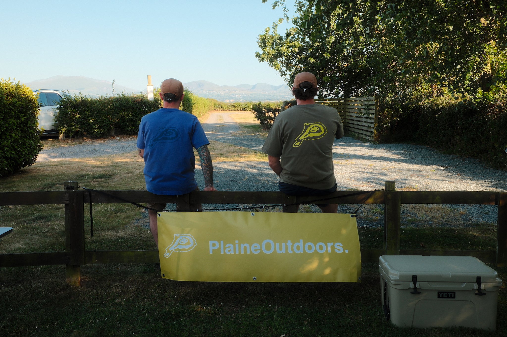 Two men standing behind a wooden fence outdoors, looking down a gravel path. A yellow banner in front of the fence reads 'PlaineOutdoors.